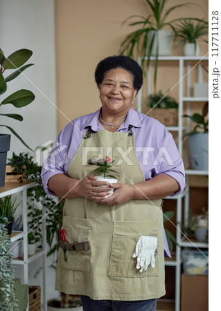 Waist up portrait of kind African American senior woman wearing apron and holding tiny plant while enjoying gardening at home 117711128