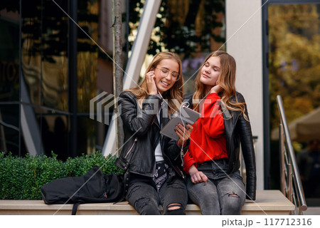 Two stylish girls sitting near shopping center and listening music of watching video on tablet. Two stylish girls sitting near shopping center and listening music of watching video on tablet. 117712316