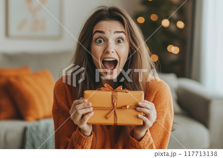 Surprised woman is holding a christmas present in her hands 117713138