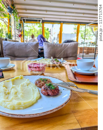 Lunch at the restaurant. A portion of beef patties in Russian style with mashed potatoes and herbs. In the background - salad with beetroot and herring, a cup of coffee and the interior of the cafe. Lunch at the restaurant. A portion of beef patties in Russian style with mashed potatoes and herbs. In the background - salad with beetroot and herring, a cup of coffee and the interior of the cafe. 117713764