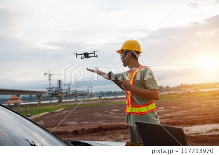 An engineer man in a yellow hard hat is standing next to a car with a drone flying above An engineer man in a yellow hard hat is standing next to a car with a drone flying above 117713970