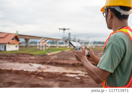 An engineer man in a yellow safety vest is holding a remote control and looking at a drone 117713971