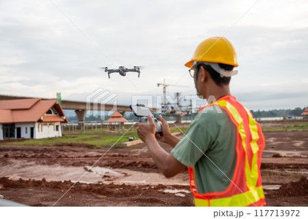 A man in a yellow safety vest is flying a drone A man in a yellow safety vest is flying a drone 117713972