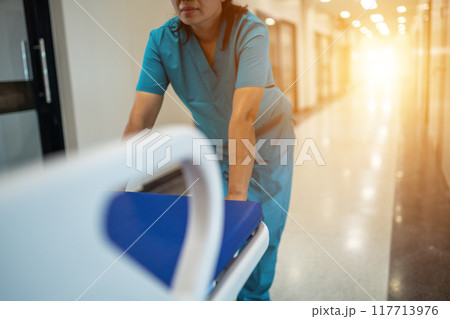 A woman in a blue scrubs is pushing a hospital bed A woman in a blue scrubs is pushing a hospital bed 117713976
