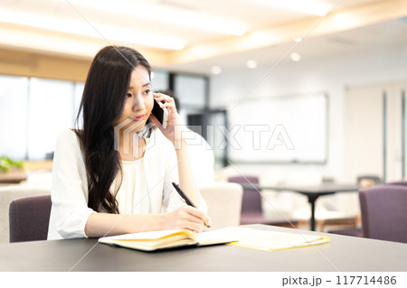 スマホの通話機能で通話しながらノートに筆記する女性 117714486