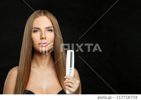Glad woman with hair straightener iron in studio isolated on black background. Haircare, wellness Glad woman with hair straightener iron in studio isolated on black background. Haircare, wellness 117716738