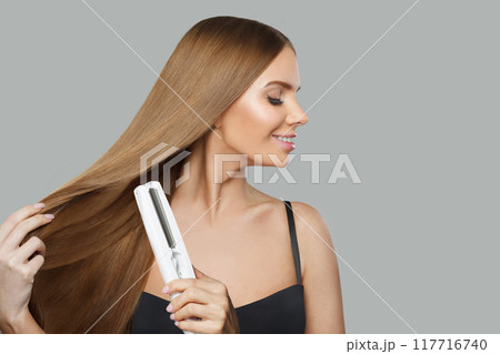 Gorgeous smiling woman with long straight healthy hairstyle using straightening iron  117716740