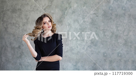 Cute young woman with curly hair and make-up in black dress posing against gray stucco plaster  117719300