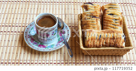 A cup of fresh black coffee and puff with berries. A cup of fresh coffee on a saucer and is fresh puff with berries lie in a straw basket A cup of fresh black coffee and puff with berries. A cup of fresh coffee on a saucer and is fresh puff with berries lie in a straw basket 117719352