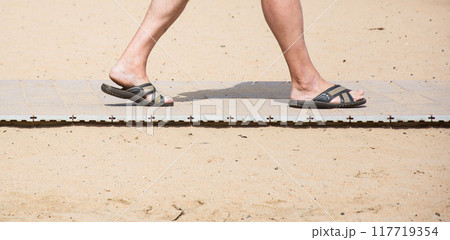 Men's feet in summer slates on the beach sand track. On the yellow sand beach lies on the path and there are feet in the summer slates Men's feet in summer slates on the beach sand track. On the yellow sand beach lies on the path and there are feet in the summer slates 117719354