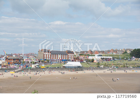遠浅の砂浜の海岸でくつろぐ人々と海岸沿いの遊園地 南ウェールズの郊外にて 遠浅の砂浜の海岸でくつろぐ人々と海岸沿いの遊園地 南ウェールズの郊外にて 117719560