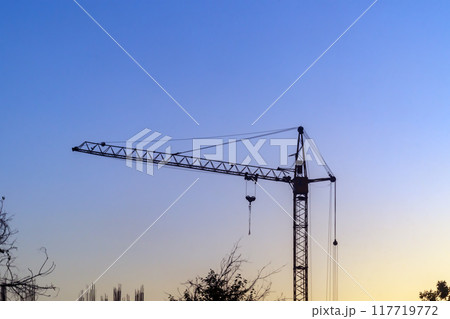 Dark silhouette of tower construction crane after work shift at sunset with soft focus against background of blue sky, yellow areola of sunset. Evening bright colors of nature Dark silhouette of tower construction crane after work shift at sunset with soft focus against background of blue sky, yellow areola of sunset. Evening bright colors of nature 117719772