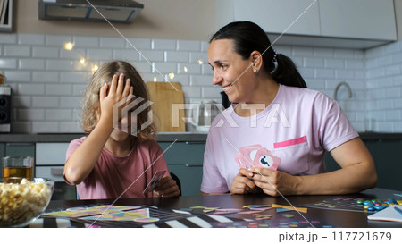 Cute daughter and her cheerful mother have fun while pulling a card from hands of each other during a kitchen board game Cute daughter and her cheerful mother have fun while pulling a card from hands of each other during a kitchen board game 117721679
