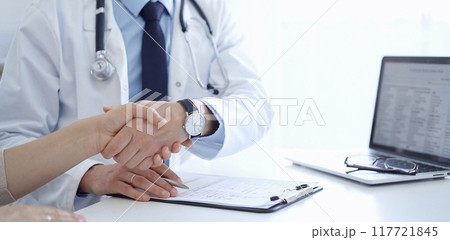 Doctor and a patient are shaking hands. The physician is wearing a white medical coat over a blue shirt. Medical service 117721845