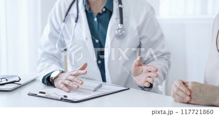 Doctor and a patient. The physician, wearing a white medical coat over a green shirt, is gesturing with his hands during a consultation in the clinic. Medicine 117721860