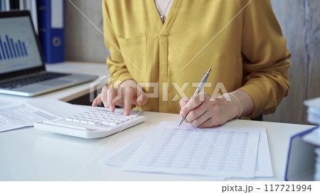 Businesswoman with yellow blues is using white calculator and taking notes in her office. Taxes and audit concept Businesswoman with yellow blues is using white calculator and taking notes in her office. Taxes and audit concept 117721994
