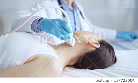 Doctor man wearing blue medical gloves is using ultrasound equipment on neck of female patient lying down for a medical examination 117722004