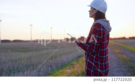 Woman engineer wearing a white protective helmet is taking notes with a tablet computer on a field with wind turbines, as the sun sets. Clean energy and engineering concept 117722020