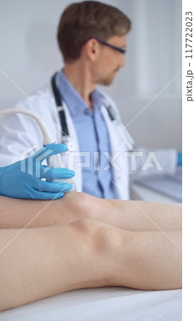 Doctor man with blue protective gloves using ultrasound equipment on the knee of a female patient, analyzing the results on a monitor in the background. Medicine Doctor man with blue protective gloves using ultrasound equipment on the knee of a female patient, analyzing the results on a monitor in the background. Medicine 117722023
