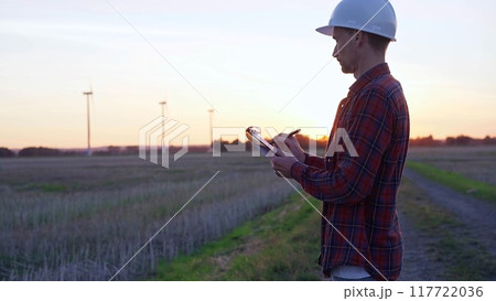 Man engineer wearing a white protective helmet is taking notes with a clipboard in a field with wind turbines, as the sun sets. Clean energy and engineering concept Man engineer wearing a white protective helmet is taking notes with a clipboard in a field with wind turbines, as the sun sets. Clean energy and engineering concept 117722036