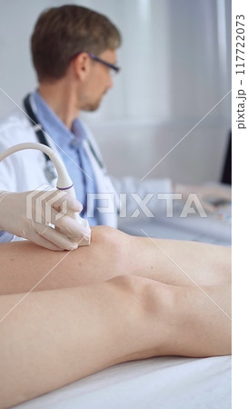 Doctor man with blue protective gloves using ultrasound equipment on the knee of a female patient, analyzing the results on a monitor in the background. Medicine Doctor man with blue protective gloves using ultrasound equipment on the knee of a female patient, analyzing the results on a monitor in the background. Medicine 117722073