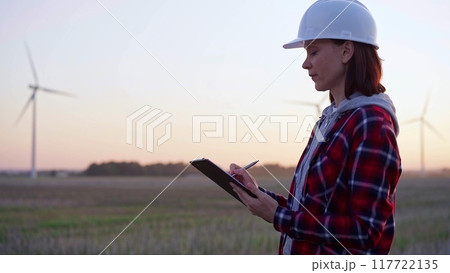 Woman engineer taking notes on a clipboard on a field with wind turbines, as the sun sets. Clean energy and engineering concept 117722135