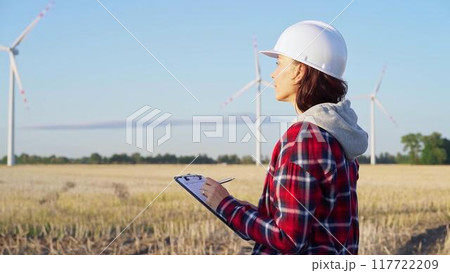 Female engineer wearing a white protective helmet is taking notes with a clipboard in a field with wind turbines, as the sun sets. Clean energy concept Female engineer wearing a white protective helmet is taking notes with a clipboard in a field with wind turbines, as the sun sets. Clean energy concept 117722209