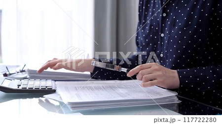 Businesswoman using calculator and reviewing ring folder of financial documents with magnifying glass at desk in modern office. Audit and taxes in business Businesswoman using calculator and reviewing ring folder of financial documents with magnifying glass at desk in modern office. Audit and taxes in business 117722280