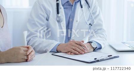 A doctor and a patient. The physician, wearing a white medical coat over a blue shirt, is filling out a medical record form during a consultation in the clinic. Medical service A doctor and a patient. The physician, wearing a white medical coat over a blue shirt, is filling out a medical record form during a consultation in the clinic. Medical service 117722340
