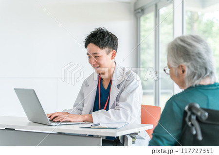 病院・介護施設で高齢者女性を笑顔で診察・問診する若いアジア人男性医師（かかりつけ医） 117727351