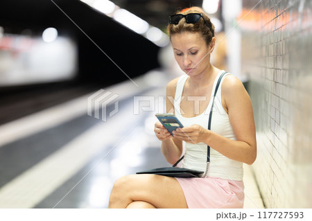 Adult woman sits at subway station with phone 117727593