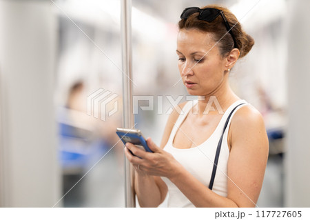 Woman standing in subway wagon, holding on handrails, browsing in smartphone Woman standing in subway wagon, holding on handrails, browsing in smartphone 117727605
