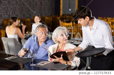 Young male waiter takes order from elderly couple in restaurant Young male waiter takes order from elderly couple in restaurant 117727633