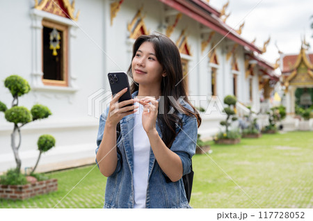 A beautiful, positive Asian female tourist strolls around a temple with a smartphone in her hand. A beautiful, positive Asian female tourist strolls around a temple with a smartphone in her hand. 117728052