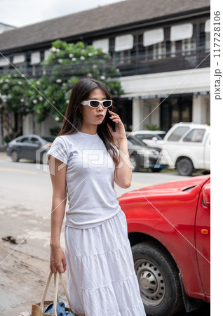 A fashionable Asian woman is talking on the phone while walking by the street in the city. 117728106