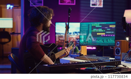 Music producer mixing guitar sounds played by him on acoustic instrument, using electronic soundboard console and daw software. Skilled musician developing new tunes, home studio. Camera A. 117730834