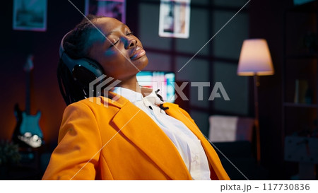 African american student relaxing with ambience music on headphones, working on new school assignment with songs in the background. Young woman enjoys distance education. Camera B. 117730836