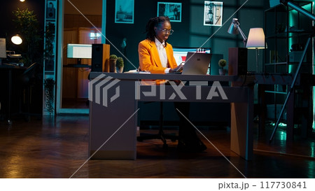 African american student works on school assignments at home, researching important information for her final bachelor essay. Young woman writing project paper at desk. Camera B. 117730841