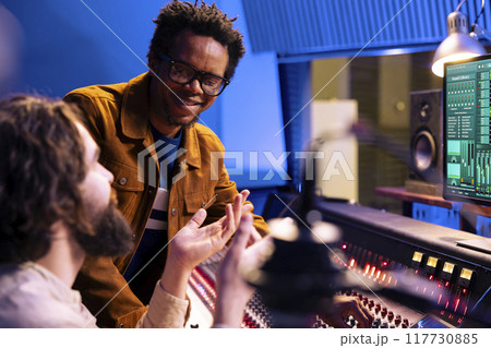 African american composer works with engineer in recording studio, talking about adding new sound effects on songs recorded. Young artist and producer editing records with control panel board. 117730885