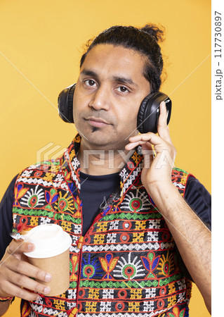 Portrait of fatigued man listening music and drinking caffeinated beverage to recover from sleepless night, suffering from insomnia. Drowsy indian person hearing songs, sipping coffee to wake up Portrait of fatigued man listening music and drinking caffeinated beverage to recover from sleepless night, suffering from insomnia. Drowsy indian person hearing songs, sipping coffee to wake up 117730897