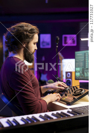 Young musician composing soundtracks and editing them on mixing console, creating cool tunes on daw software interface in his home studio. Producer adjusting volume levels on stereo gear. Young musician composing soundtracks and editing them on mixing console, creating cool tunes on daw software interface in his home studio. Producer adjusting volume levels on stereo gear. 117731027