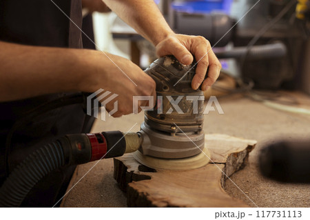 Artisan in studio enjoying diy hobby, using orbital sander with fine sandpaper on lumber to achieve smooth finish. Creative person in carpentry shop uses angle grinder to create wooden objects 117731113