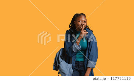 Stern african american woman doing shushing hand gesturing, irritated by noise, having negative mood. Girl placing finger on lips, doing quiet sign gesture, studio background, camera B 117731119