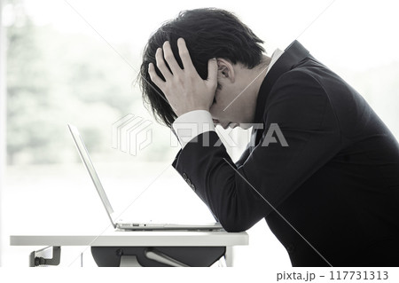デスクで頭を抱えながら悩む若い男性ビジネスマン デスクで頭を抱えながら悩む若い男性ビジネスマン 117731313