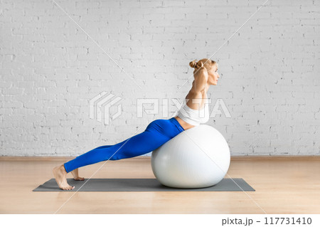 Fit Caucasian woman practice back extension exercise with hands behind her head using fitness ball in loft studio indoor. 117731410