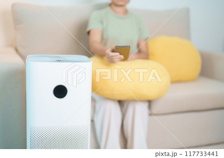 Woman turn on Air Purifier in living room by smartphone application. Purification system for filter and cleaning dust PM2.5 HEPA and virus. Allergy, Pure air, health lifestyle and Air Pollution Woman turn on Air Purifier in living room by smartphone application. Purification system for filter and cleaning dust PM2.5 HEPA and virus. Allergy, Pure air, health lifestyle and Air Pollution 117733441
