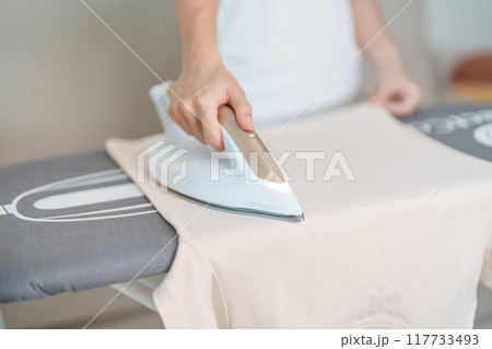Woman hold Electric hot iron press T shirt clothes on ironing board, Intelligent automatic steam iron. Laundry, Housework, Housekeeping, Domestic hygiene, household of chores and daily routine concept 117733493