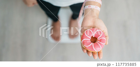 woman on weight scale and hand hold pink Donut, choose stop eating sweet is Unhealthy ealthy food. Dieting control, Weight loss, Obesity, eating lifestyle and nutrition concepts woman on weight scale and hand hold pink Donut, choose stop eating sweet is Unhealthy ealthy food. Dieting control, Weight loss, Obesity, eating lifestyle and nutrition concepts 117735885