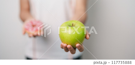 woman hand hold green Apple and donut, female fitness choose between fruit is Healthy and sweet is Unhealthy junk food. Dieting control, Weight loss, Obesity, eating lifestyle and nutrition concept 117735888