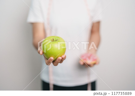 woman hand hold green Apple and donut, female fitness choose between fruit is Healthy and sweet is Unhealthy junk food. Dieting control, Weight loss, Obesity, eating lifestyle and nutrition concept 117735889